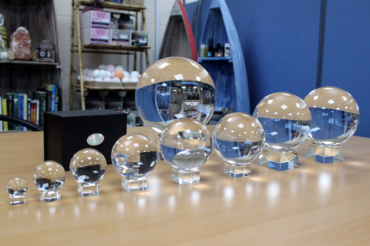 Image shows a selection of crystal balls in a row ranging from smallest, 30mm to largest at 150mm. On a table with shelving and books behind  a typical shop layout