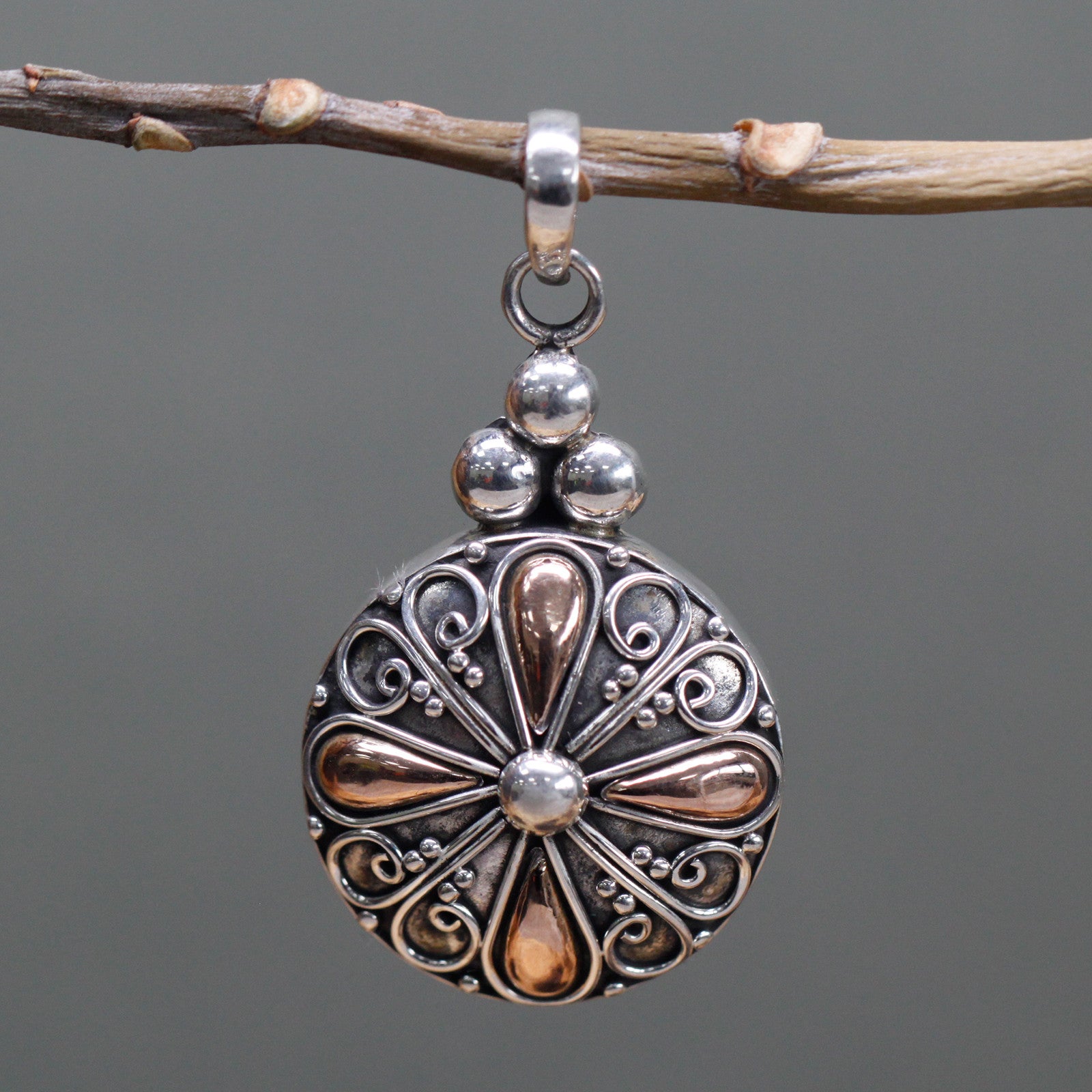 Image of a classic round pendant crafted from silver and gold, handcrafted in Indonesia. Image shows pendant close up hanging off a  twig like object