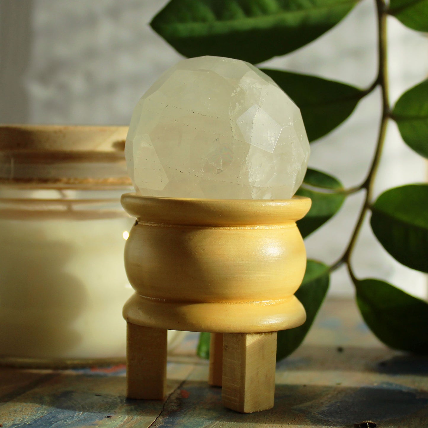 Gemstone Faceted Healing Ball & Stand - Rock Quartz. Image of a Rock Quartz Gemstone Healing Ball accompanied by a wooden stand, perfect for energy amplification and spiritual practices. Background has a green plant and jar on wooden table top