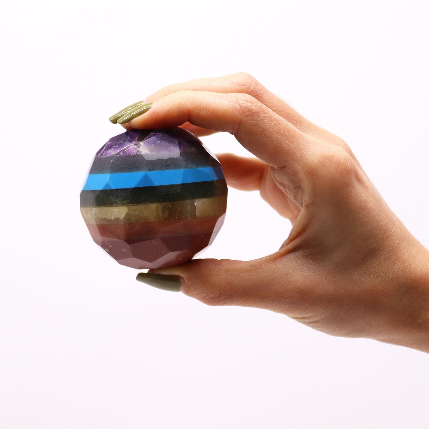 Image of a hand holding in the fingers the gemstone faceted healing ball, 7 chakrs colours against white background. Shows the sizing of the ball in contrast to human hand. 