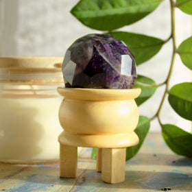 Gemstone Faceted Healing Ball & Stand - Amethyst. Image shows the gemstone on the 3 legged wooden stand, on a wooden table adorned with green leaves and glass jar for ambience