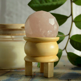 Gemstone Faceted Healing Ball & Stand - Rose Quartz. Image shows the gemstone healing ball on the wooden 3 legged stand on a wooden top also with green leaves and glass jar in background for ambience.